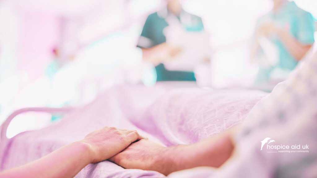 one hand holding the hand of a patient in bed in a hospice, while two nurses are blurred in the background