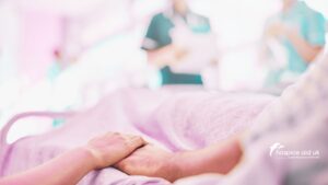one hand holding the hand of a patient in bed in a hospice, while two nurses are blurred in the background