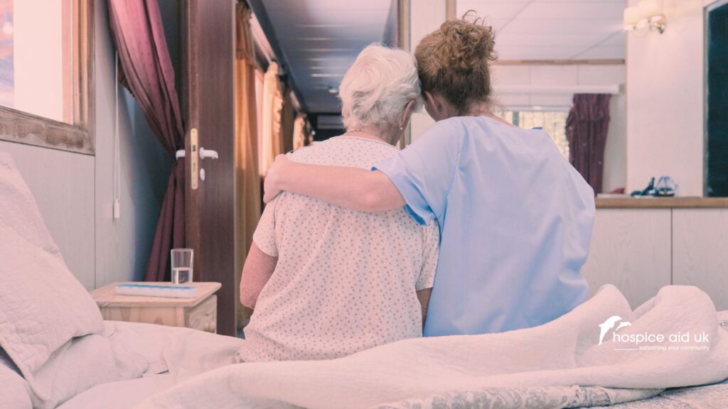 two women have their backs to camera whilst sitting together on a hospice bed. One is a patient with a terminal illness, the other is her hospice nurse who has her arm around the other woman.