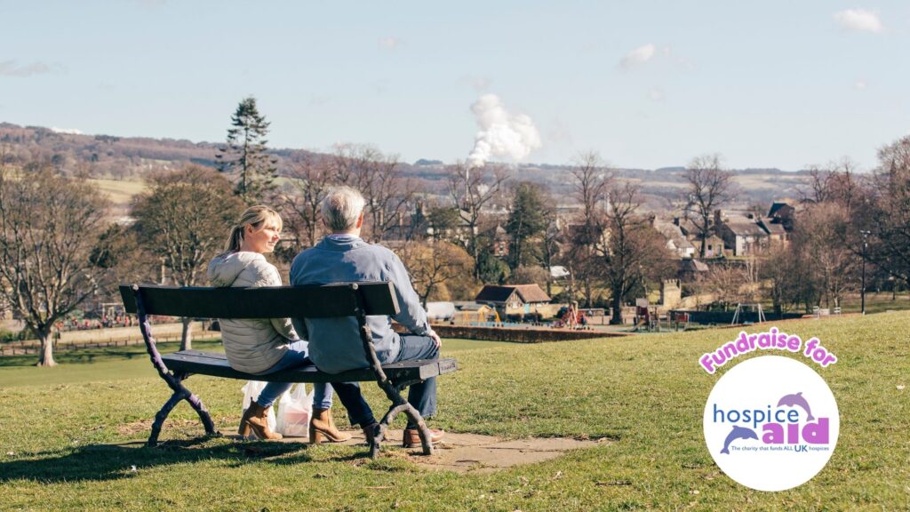 A young woman sits beside a mature man on a park bench in the countryside, overlooking a village scene.